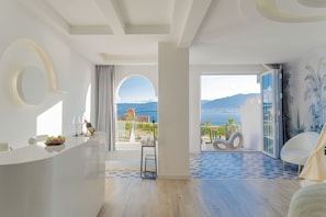 Una moderna camera d'hotel con una reception dalla forma curva, vista sul mare e sulle montagne, un balcone con pavimento decorato e una sedia bianca.