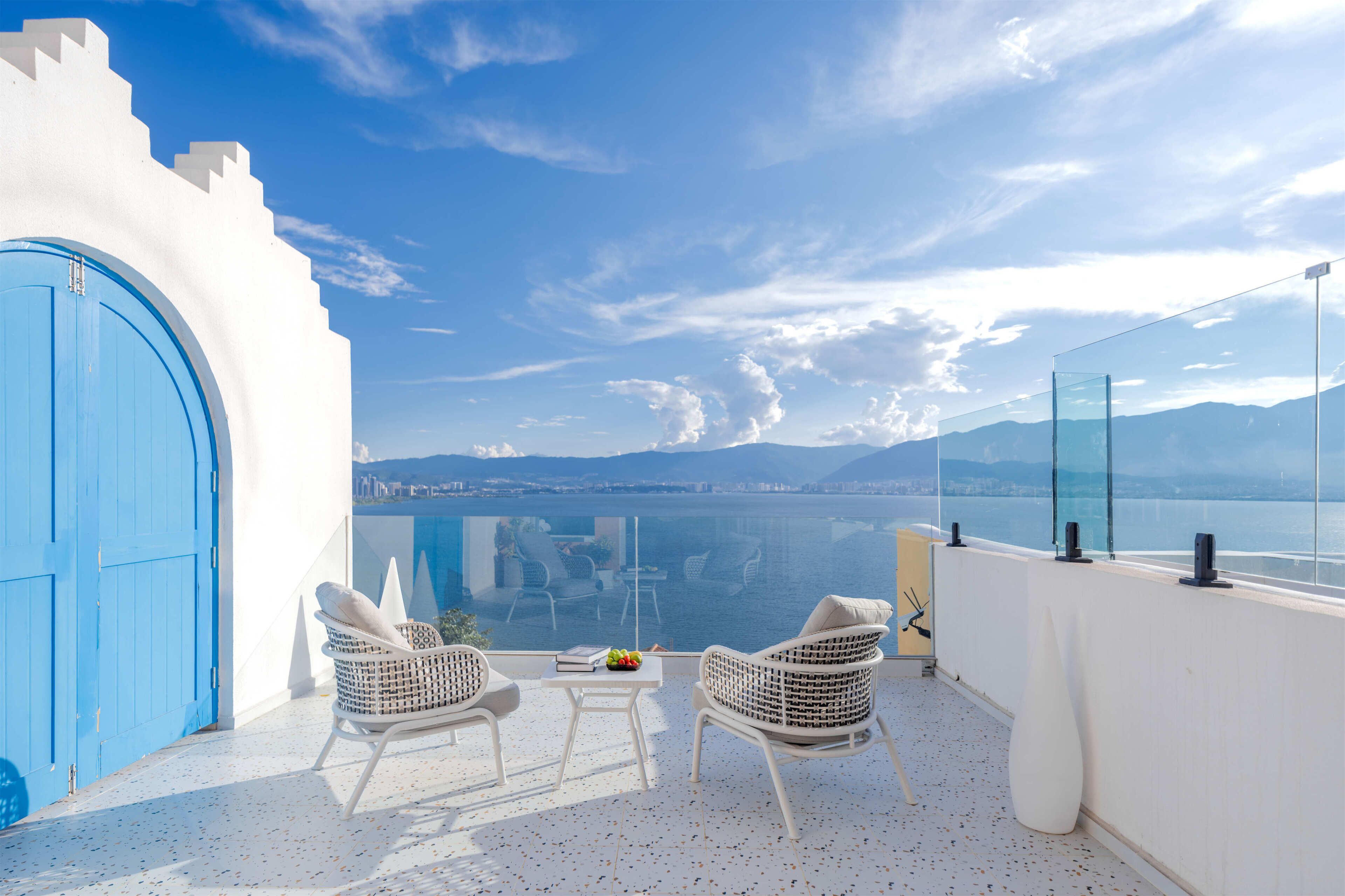 A balcony with two wicker chairs, a small table with a book and fruit, and a view of a city and sea.