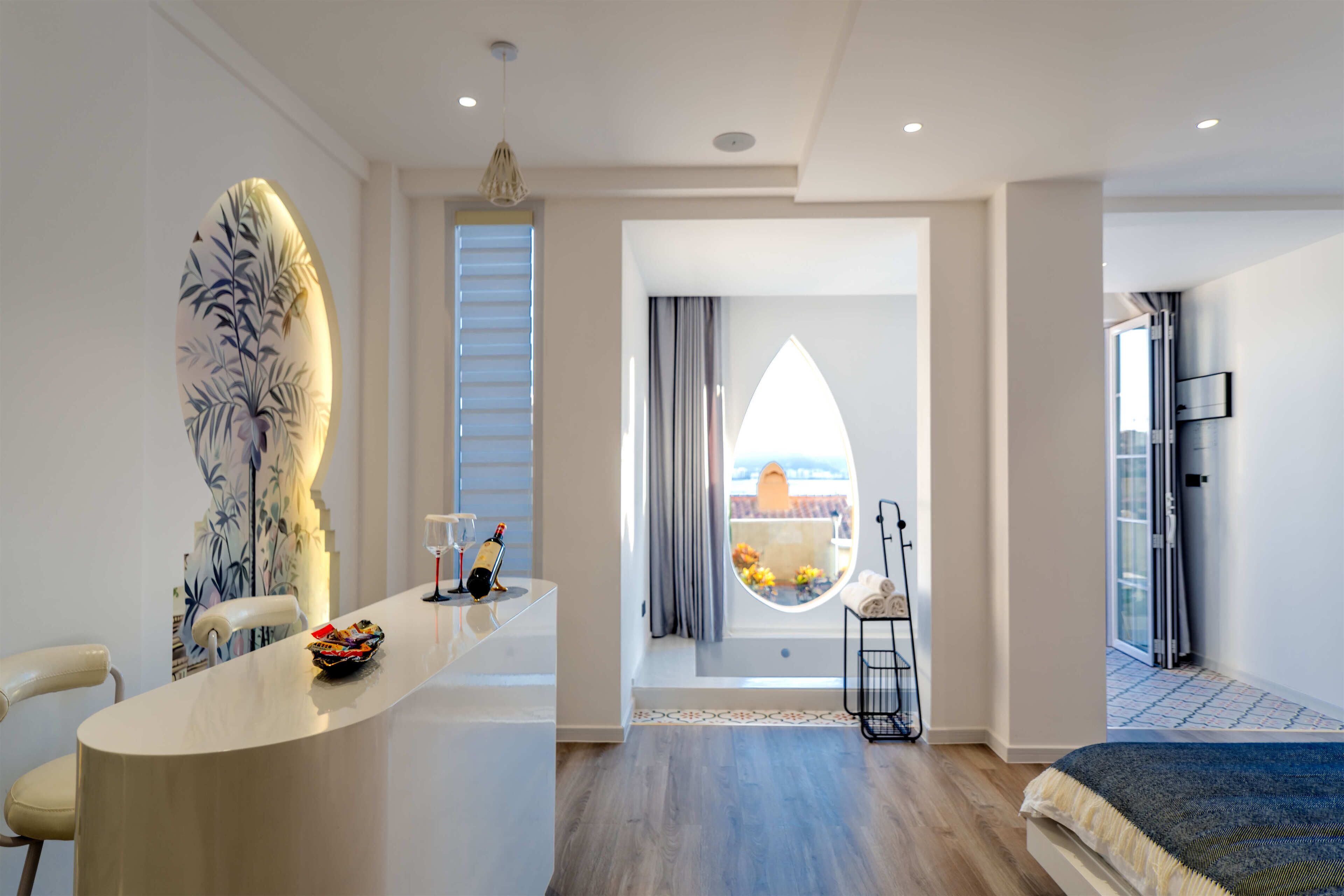 A modern hotel room with a large window, a bed, a seating area with a table, and a decorative wall piece.