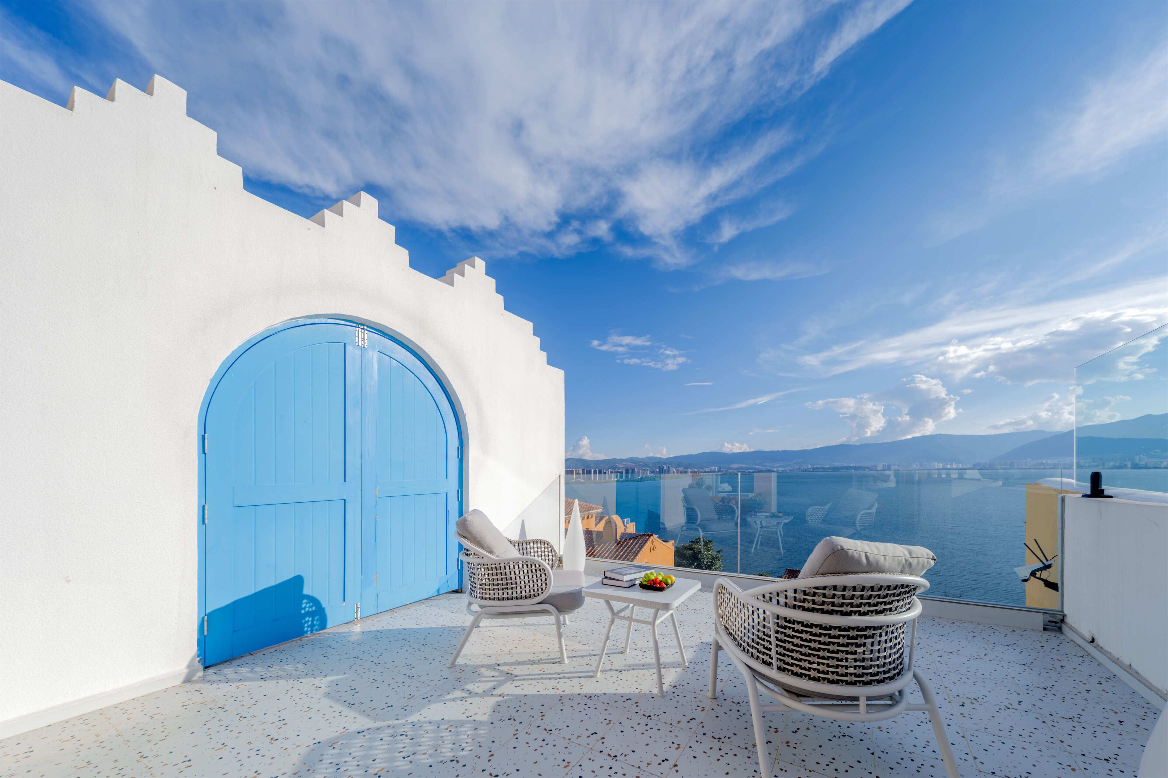 A balcony with a white wall, a blue door, and a view of the sea and cityscape.