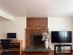A living room with a brick fireplace, a wooden desk, a black office chair, a white vase with flowers, and a flat-screen TV on a glass stand.