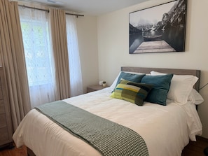 Una cama bien hecha con una colcha estampada y almohadas, una ventana con cortinas y un cuadro en la pared.
