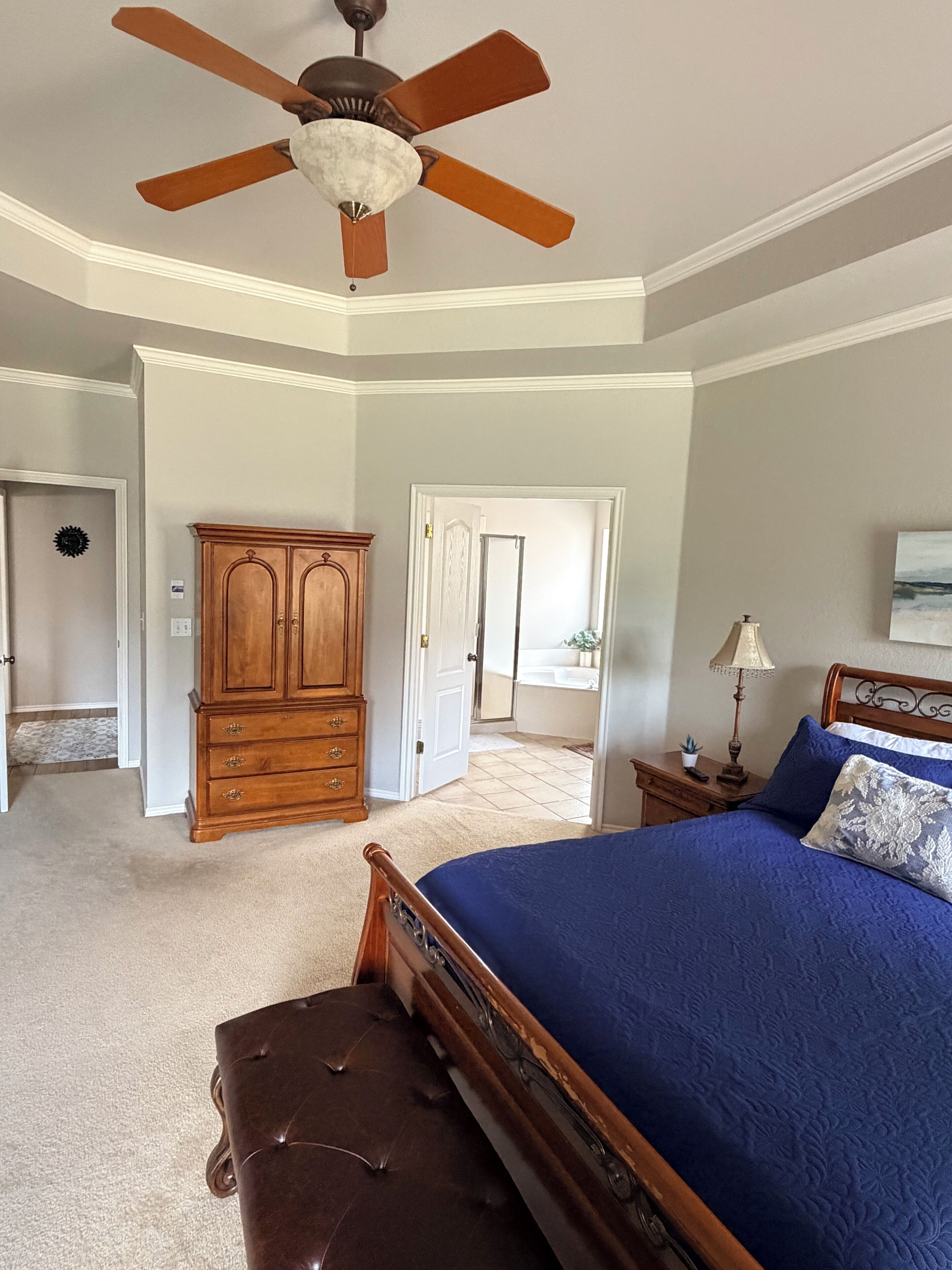 A bedroom with a bed, a wooden dresser, a ceiling fan, and a door leading to a bathroom.