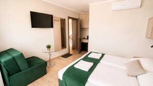 Een hotelkamer met een bed, een groene bank, een kleine tafel met een plant, een aan de muur bevestigde televisie en een keukenruimte zichtbaar door een open deur.