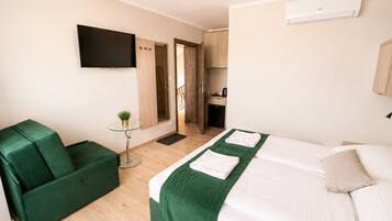 A hotel room with a bed, a green sofa, a small table with a plant, a TV mounted on the wall, and a kitchen area visible through an open door.