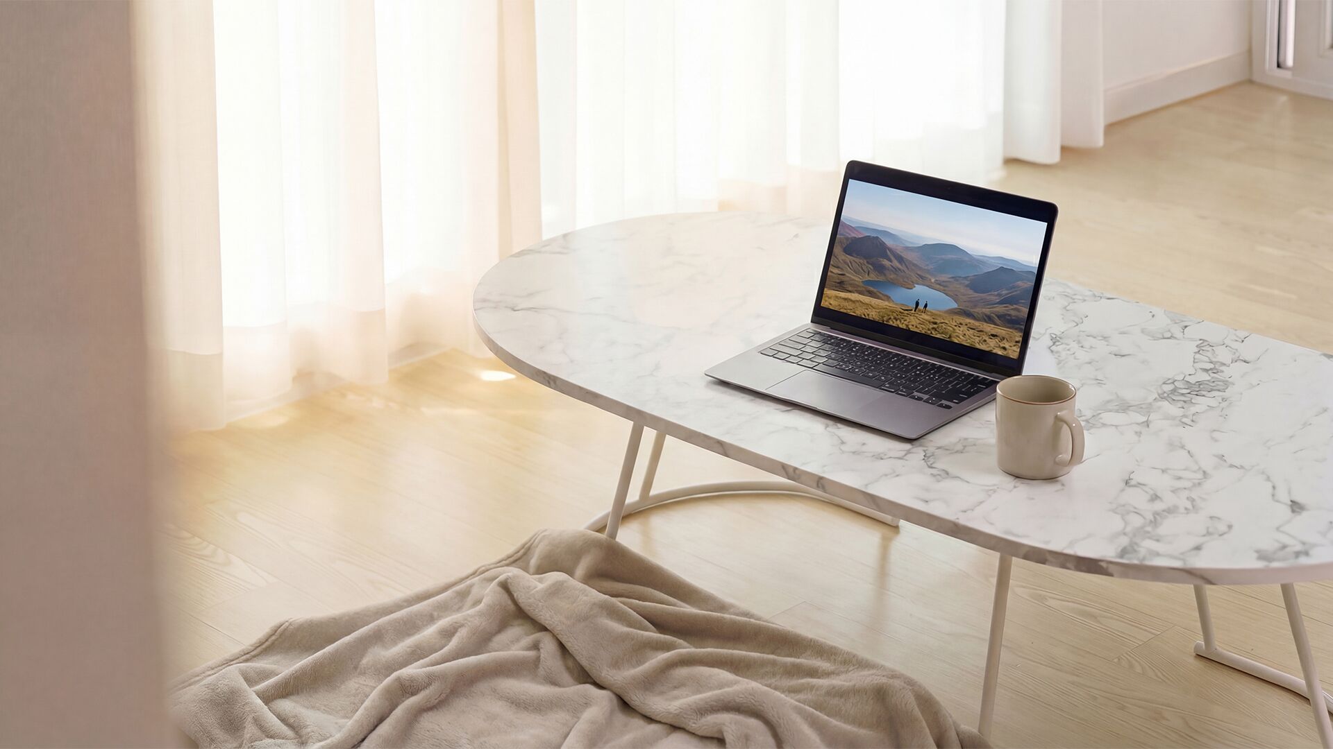 A laptop on a marble table with a mountain view on the screen, a mug, and a blanket on the floor.