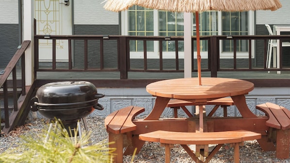 A wooden picnic table with a thatched umbrella, a barbecue grill, and a railing with a view of a building.