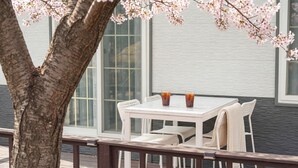 A white outdoor table with two drinks, surrounded by white chairs, set against a building with large windows and a tree with pink blossoms. - Daebudo Blue Island Camping Site (Ansan)
