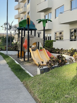 Ein Spielplatz mit Rutschen und Klettergerüsten, umgeben von einer angelegten Grünfläche und einem Gebäude mit Balkonen.