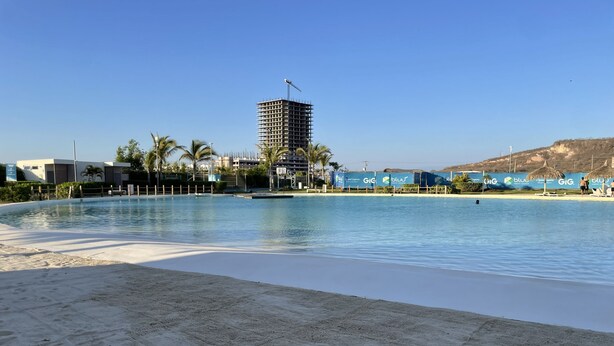 Une piscine avec une zone de plage de sable, un grand immeuble en arrière-plan et des palmiers.