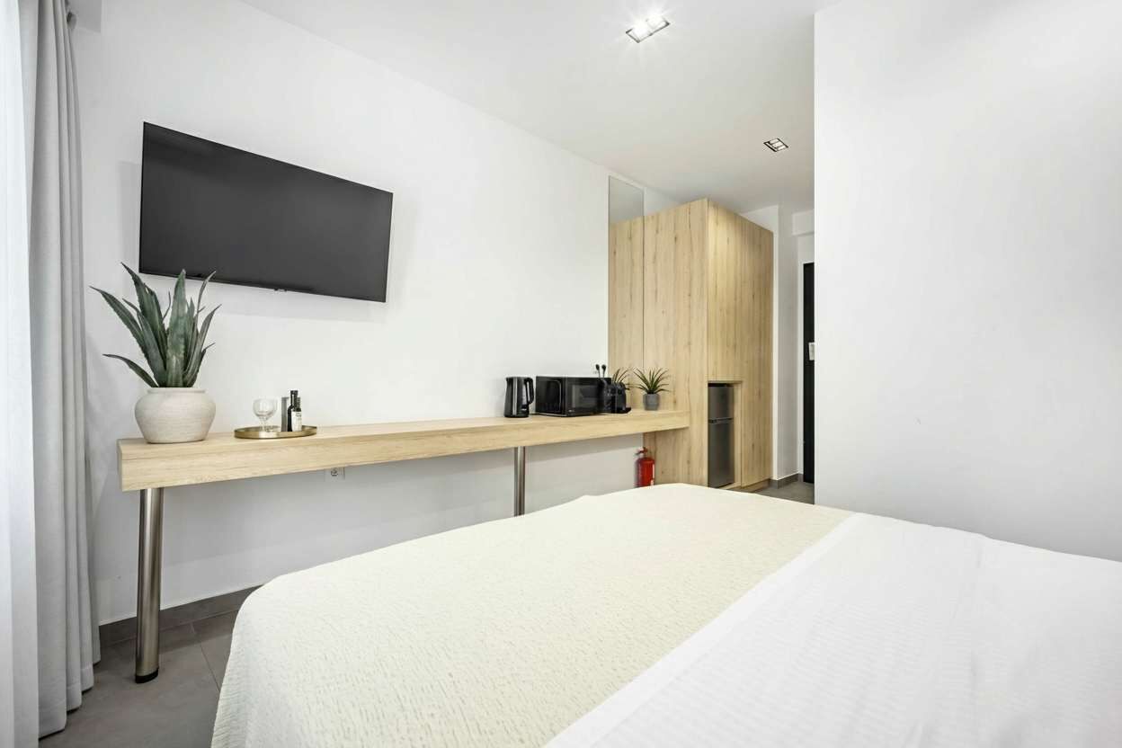 Ein modernes Hotelzimmer mit einem Bett, einem Wand-Fernseher, einem Holztisch mit Pflanze, einem Wasserkocher und einer Mikrowelle.