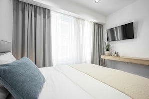 A modern hotel room with a bed, curtains, a mounted TV, and a bedside plant. - Cozy Studio Syntagma Worn Bldg Entrance (Athina)