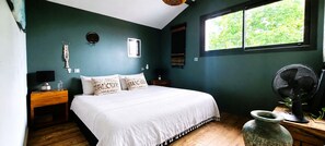 Une chambre à coucher comprenant un lit, une fenêtre, un ventilateur fixé au mur, une table de chevet et un vase décoratif.