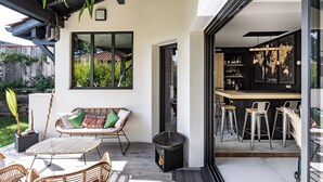 Ein Außenbereich mit Korbmöbeln, einer Bar mit Hockern und einem großen Fenster mit Blick auf einen Garten.