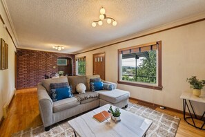 A living room with a sectional sofa, a coffee table, a brick accent wall, a large window with a view, and a ceiling light fixture.