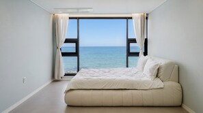 A bedroom with a bed, a large window with curtains, and a view of the sea. - Pohang Pine Wave Pension (Pohang)