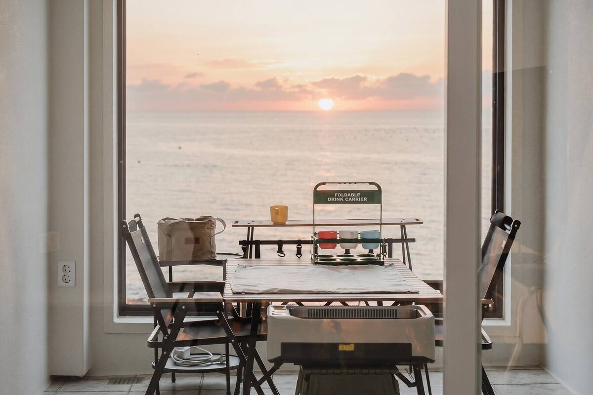Una habitación con vista al mar al atardecer, una mesa con un organizador para bebidas y una silla.
