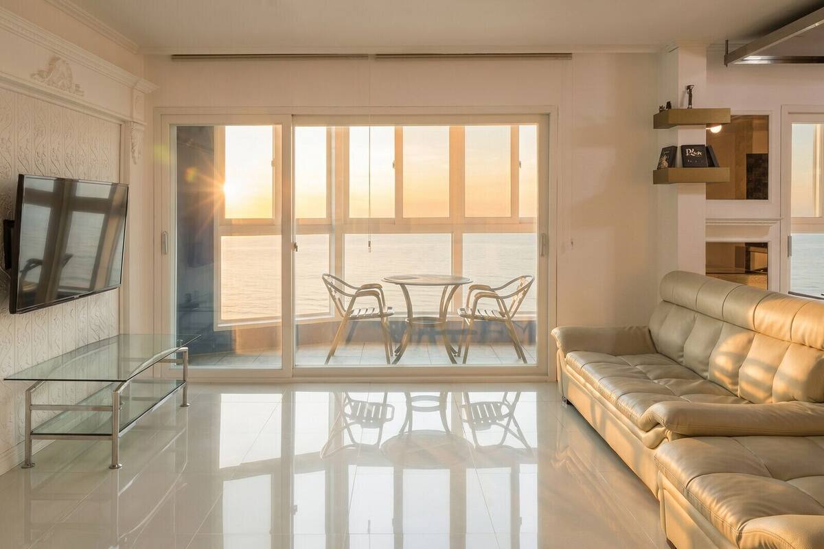 Una sala de estar moderna con una mesa y sillas de vidrio, un sofá de cuero, un televisor montado y vista al mar a través de amplias puertas de cristal.