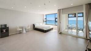 A modern bedroom with a bed, a dining table, chairs, a TV, and a view of the sea.
