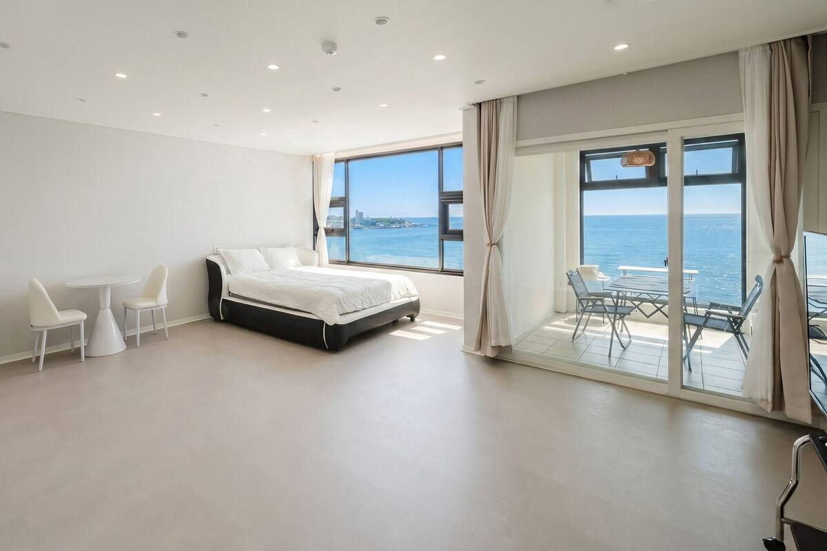 Un dormitorio moderno con cama, una mesa de comedor, sillas, televisor y vista al mar.