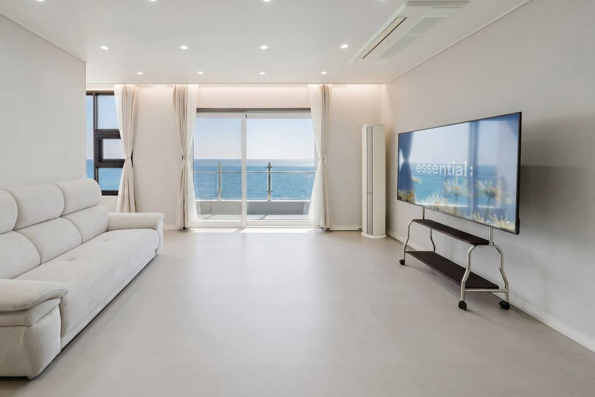 Una sala de estar moderna con un sofá, un televisor sobre un mueble bajo y un ventanal con vistas al mar.