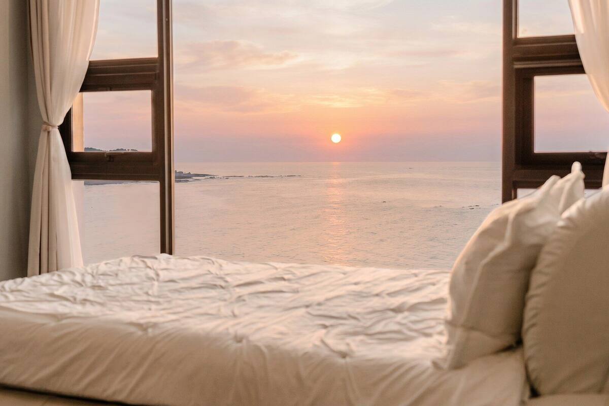 Una cama con vista al mar y al atardecer a través de una ventana.