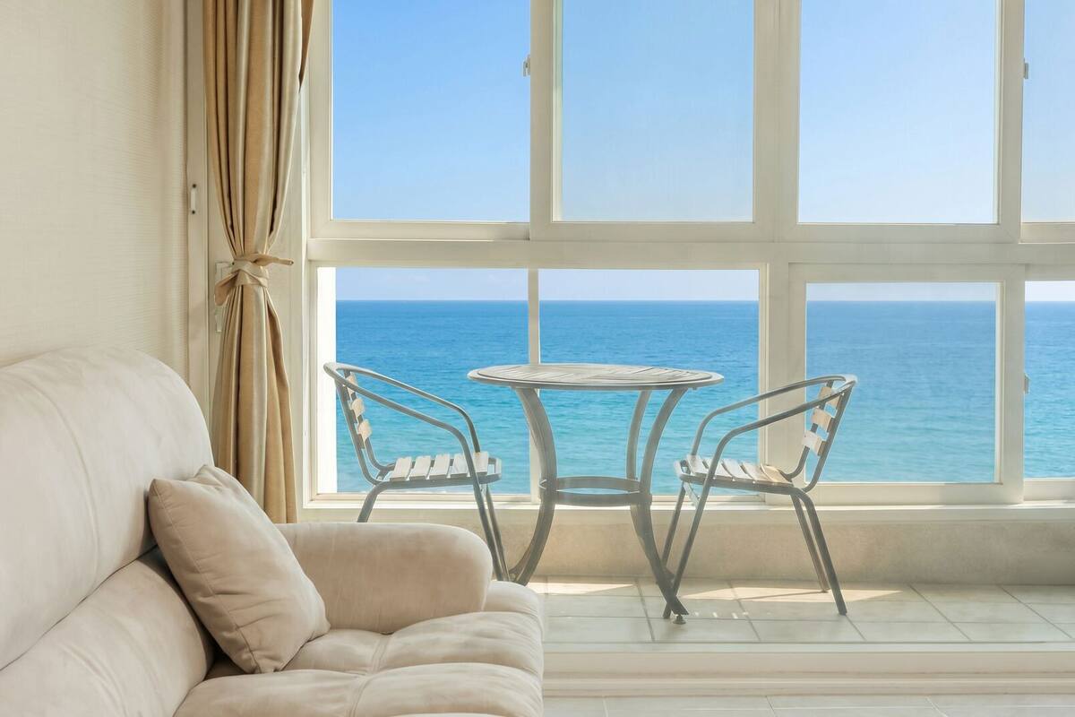 Una habitación con un sofá blanco, una mesita redonda pequeña y dos sillas frente a un ventanal con vista al océano.