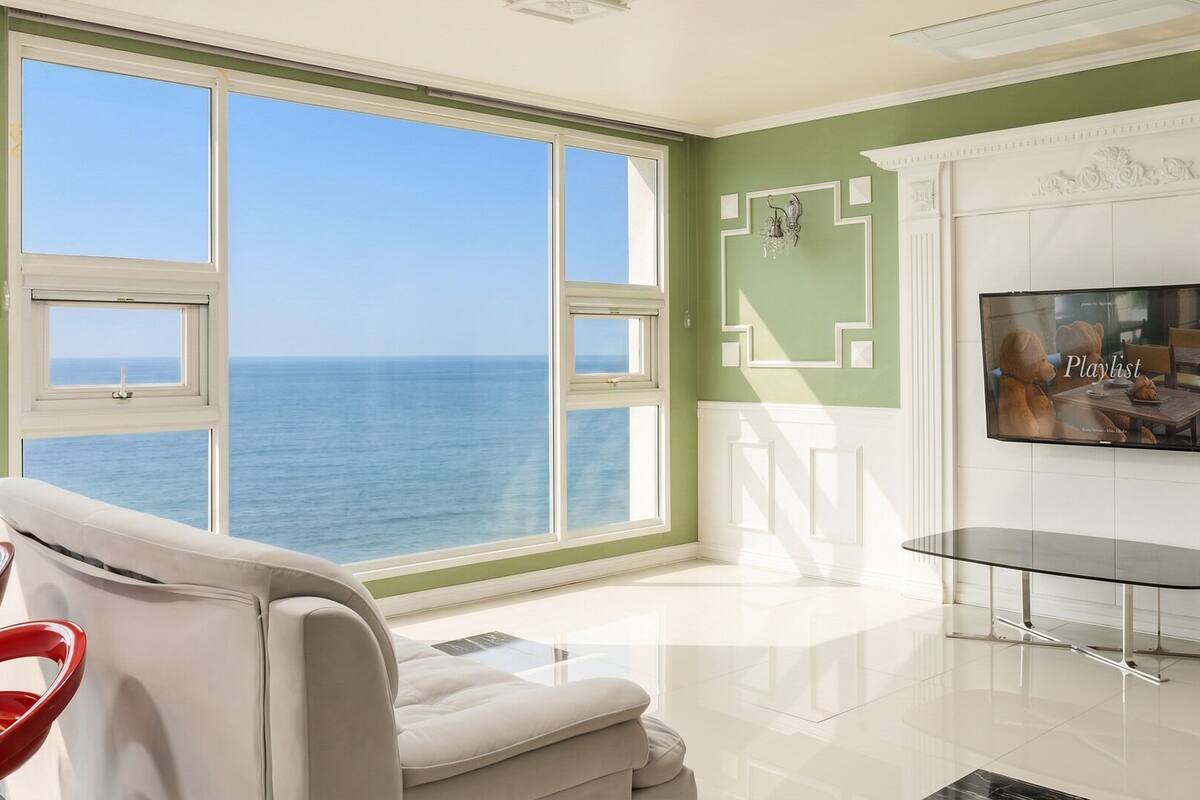 Una habitación con un amplio ventanal con vista al mar, un televisor de pantalla plana en la pared y una mesa de centro de vidrio.