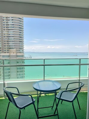 Uma varanda com guarda-corpo de vidro, duas cadeiras, uma mesa redonda pequena e vista para um prédio e uma praia.