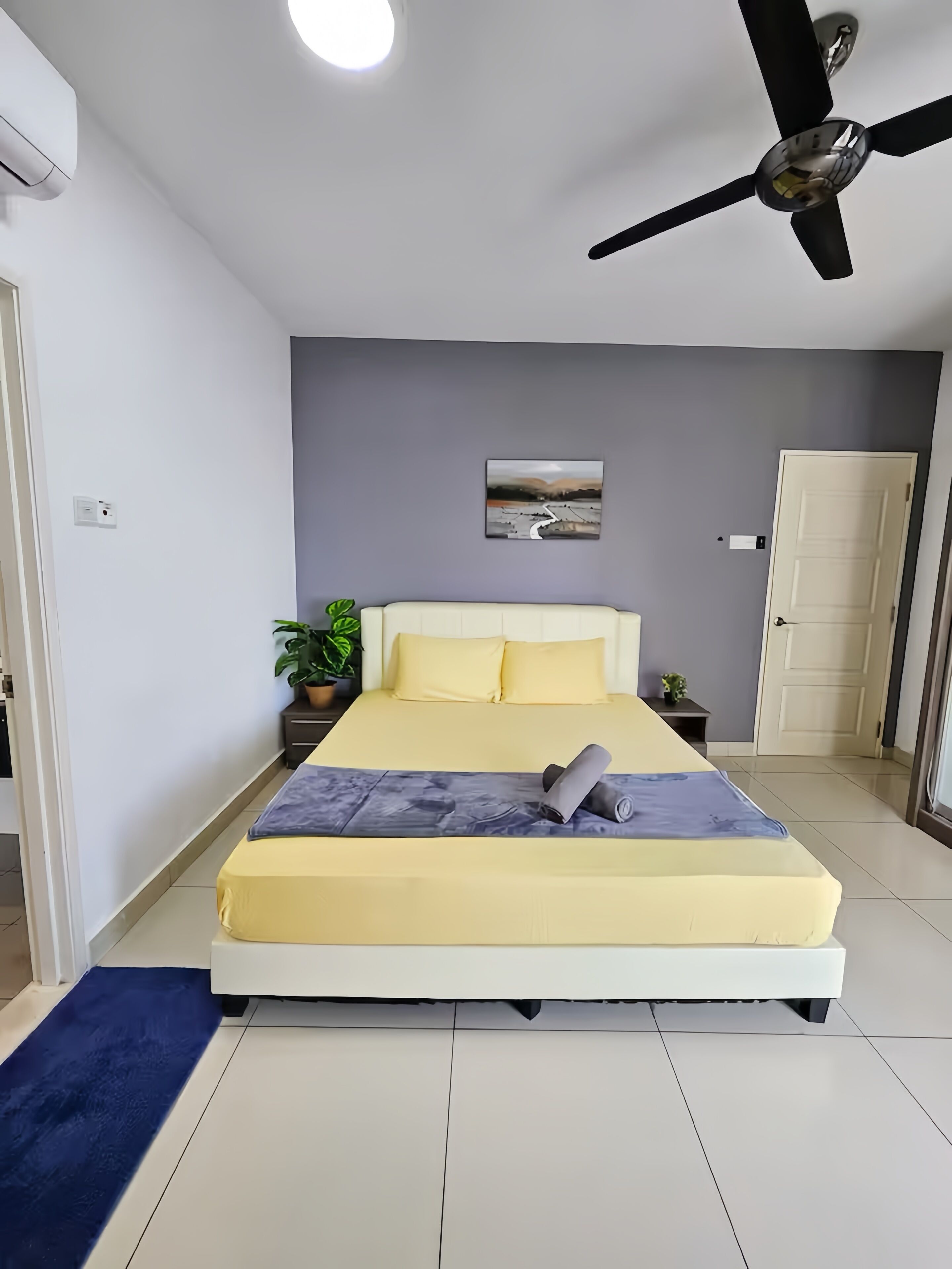 Quarto com cama, ventoinha no teto, quadro na parede, planta em vaso e porta.