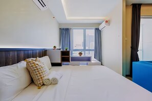 Habitación de hotel con cama, un pequeño rincón de descanso con una planta y vistas a edificios a través de amplias ventanas.