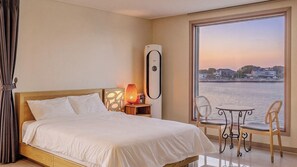 A hotel room with a bed, a heater, a table with two chairs, and a view of a body of water.