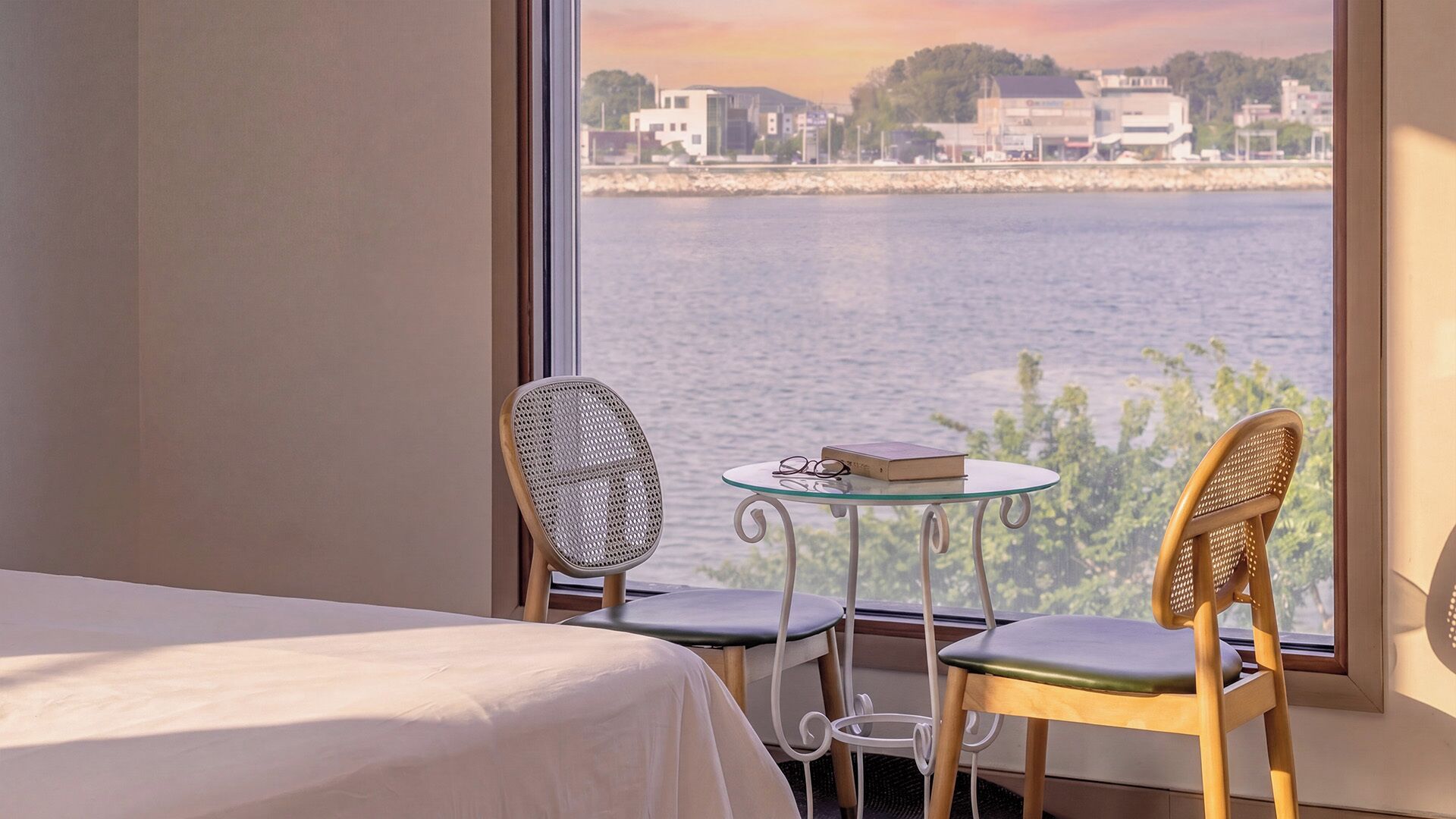 A room with a bed, two chairs, a small table, and a view of a body of water with buildings in the distance.