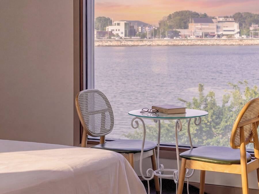 A room with a bed, two chairs, a small table, and a view of a body of water with buildings in the distance.