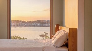 Una cama con cabecera de madera, vista a un paisaje urbano a través de un cuerpo de agua, y una ventana con vistas al atardecer.