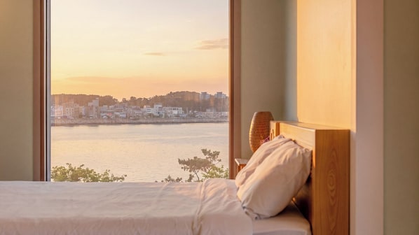 A bed with a wooden headboard, a view of a cityscape across a body of water, and a window with a view of the sunset.