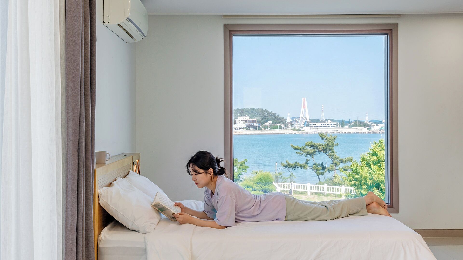 A room with a bed, a window with a view of a bridge and water, and an air conditioning unit.