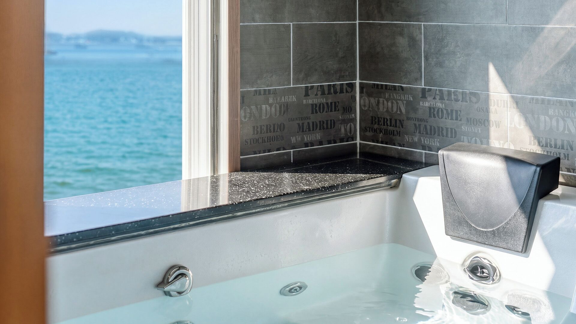 A bathroom with a bathtub, a window with a view of the sea, and a tiled wall with city names.