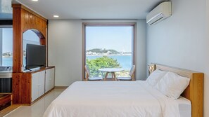 A hotel room with a bed, a TV on a wooden cabinet, a table with chairs, and a large window with a view of the sea and buildings.