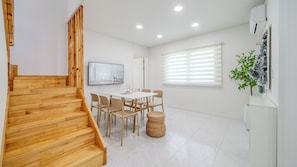 Un interior moderno con una escalera de madera, un comedor con sillas y una mesa, un ventanal con persianas y un aire acondicionado fijado a la pared.
