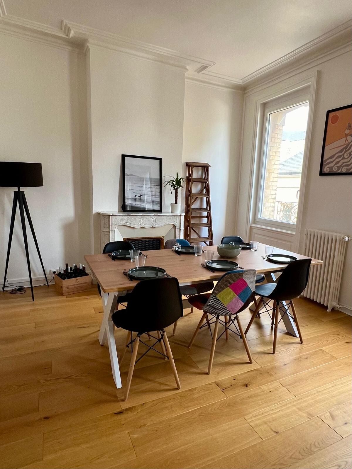 A dining room with a wooden table set for six, black chairs, a white fireplace, a framed picture, and a window with a view.