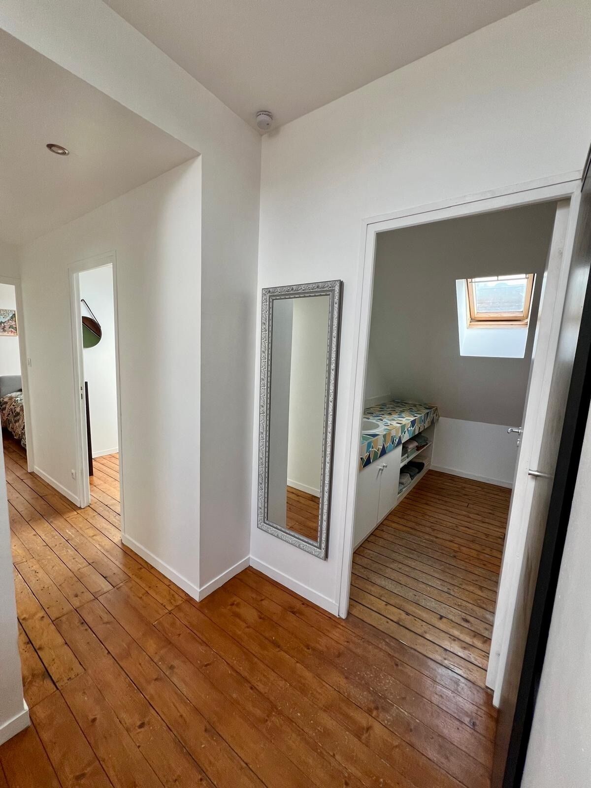 A room with wooden flooring, a large mirror, a small window, and a door leading to another room.