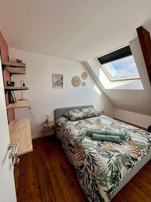 Une chambre avec un plafond en pente, un puits de lumière, un lit recouvert d’une couvre-lit à motifs de feuilles, une table de chevet et une étagère fixée au mur.
