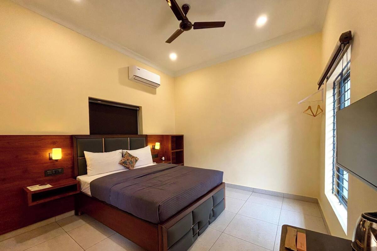 Habitación de hotel con cama, ventilador de techo, aire acondicionado, mesita de noche y televisor. Cuenta también con una ventana con persianas.