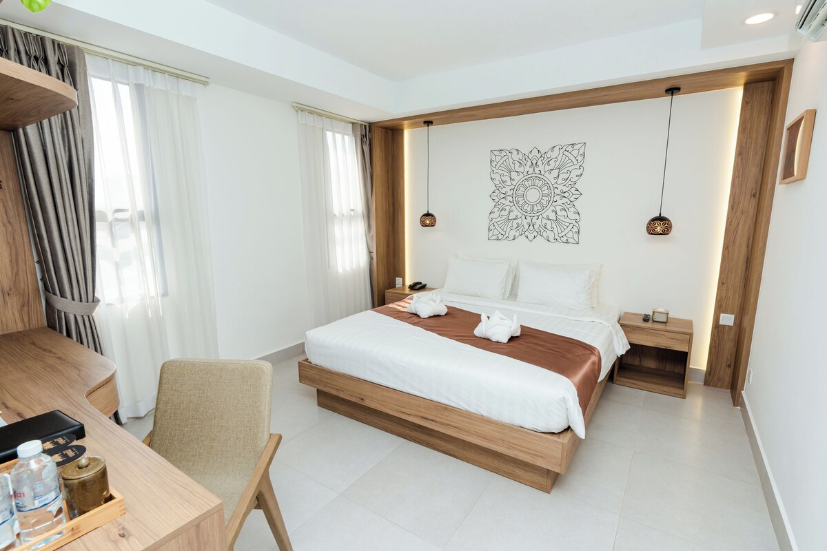 A modern hotel room with a wooden bed, a desk with a chair, a bedside table, and a wall-mounted artwork.