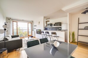 Ein modernes Wohnzimmer mit Sofa, Esstisch, Stühlen, einer Küchenzeile mit Geräten und großen Fenstern.