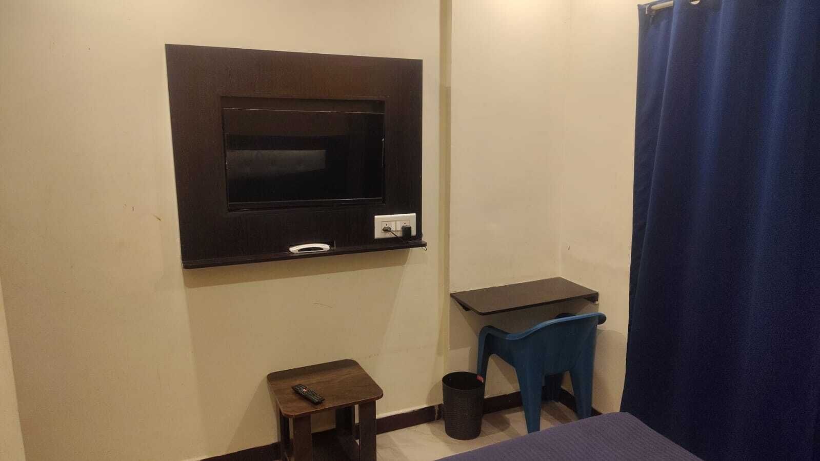 Een kleine kamer met een aan de muur bevestigde televisie, een tafeltje, een stoel en een gordijn.