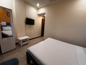 Quarto de hotel com uma cama, uma pequena mesa, um banco, uma televisão fixada na parede e uma porta que dá para outro cômodo.