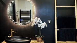 Un baño moderno con un espejo redondo, un lavabo negro, tocador de madera, orquídeas blancas y un perchero con toallas.