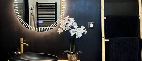 A modern bathroom with a round mirror, black sink, wooden vanity, white orchids, and a towel rack with towels.